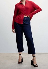 Chemise rouge à manches longues avec des boutons, assortie à un jean raccourci droit bleu foncé. Des sandales à talons carrés noires accentuent la tenue.
