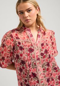 Blusa floreale in delicato tessuto rosa trasparente con fiori rossi e viola, caratterizzata da una chiusura frontale con bottoni, pizzo decorativo e dettagli plissettati sulle maniche.