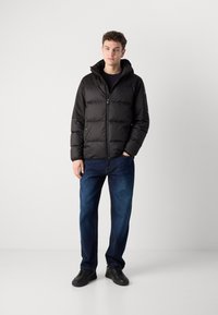 Zwarte pufferjack met een capuchon, doorgestikt ontwerp en een rits aan de voorkant. Gecombineerd met blauwe spijkerbroeken en zwarte schoenen. Minimalistische stijl.