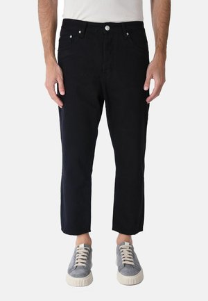 Uomo con jeans neri corti e sneakers grigie con stringhe, in piedi su sfondo bianco.