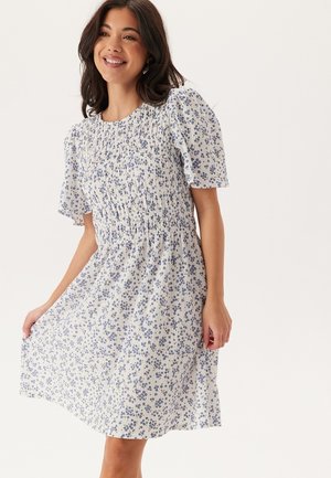 Bloemenjurk in het wit met blauwe bloemen, met een gerimpelde buste en korte pofmouwen. Gladde stof met een vloeiende rok.