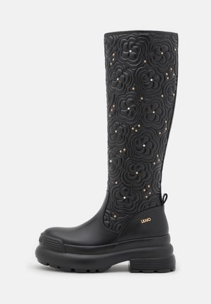 Botte noire haute jusqu'au genou avec motif floral matelassé et petits clous dorés et argentés, semelle épaisse et logo Liu Jo doré sur le talon.