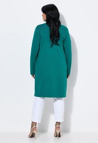 Tunic lunga color teal con taglio dritto, abbinata a pantaloni bianchi. Indossata con décolleté beige con tacco. Tessuto liscio, senza motivi.