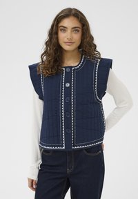 Gilet trapuntato blu navy con spalle larghe, caratterizzato da cuciture argento, sei grandi bottoni e taglio dritto. Indossato sopra una camicia leggera a maniche lunghe.