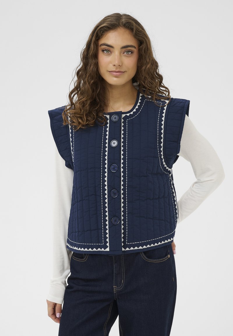 Gilet trapuntato blu navy con spalle larghe, caratterizzato da cuciture argento, sei grandi bottoni e taglio dritto. Indossato sopra una camicia leggera a maniche lunghe.