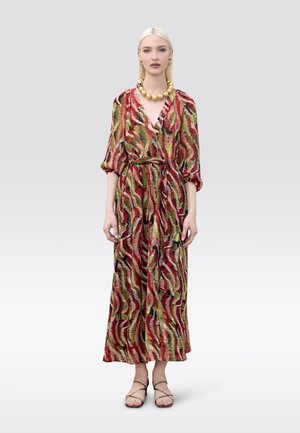 Mujer con un vestido largo de colores múltiples y estampado, con mangas abullonadas, collar de cuentas doradas, pendientes y sandalias con tiras, de pie frente a un fondo liso.