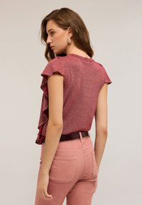 Una joven de lado lleva una blusa corta de manga con volantes roja, pantalones rosa de talle alto, un cinturón oscuro y pendientes de aro dorados.