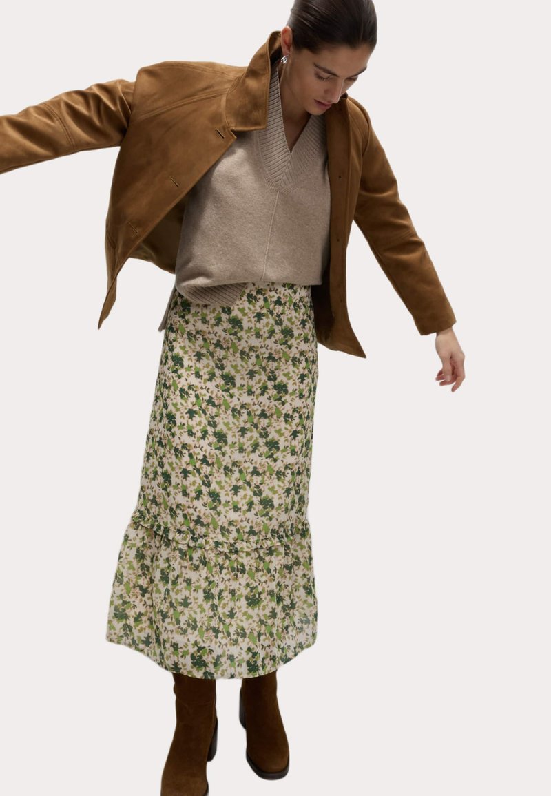 Veste en daim marron, pull en tricot beige, jupe midi florale aux tons verts et beiges, ourlet à volants superposés, et bottines brunes.