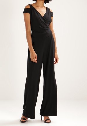 Frau trägt einen schwarzen Cold-Shoulder-Jumpsuit mit weiten Beinen und schwarzen offenen High Heels, stehend vor einem schlichten hellen Hintergrund.
