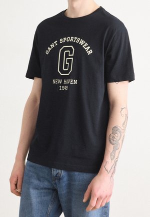 Person trägt ein schwarzes GANT Sportswear T-Shirt mit der Aufschrift „New Haven 1949“, blaue Jeans und ein Schlangen-Tattoo am linken Unterarm.