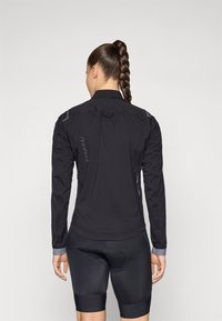 Svart sykkeljakke med lange ermer, teksturert stoff og reflekser; kombinert med svarte polstrede sykkelshorts.