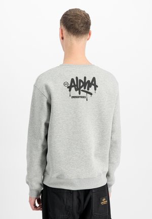 Man wearing light grey sweatshirt with "Alpha Industries" black graffiti logo on back, paired with black pants with yellow patch.