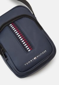 Navy modra najlon torba za čez ramo z zadrgo, ki ima dizajn z rdečimi, belimi in modrimi črtami ter logotipom "TOMMY HILFIGER" na dnu.