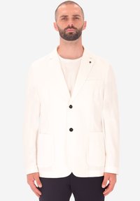 Uomo con barba che indossa un blazer bianco con due bottoni neri, una camicia bianca e pantaloni scuri, in piedi davanti a uno sfondo chiaro e uniforme.