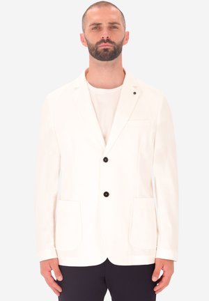 Uomo con barba che indossa un blazer bianco con due bottoni neri, una camicia bianca e pantaloni scuri, in piedi davanti a uno sfondo chiaro e uniforme.