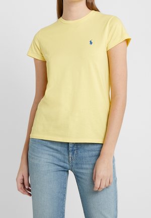 Osoba ubrana w bladożółty t-shirt z krótkim rękawem z małym niebieskim wyszywanym logo na górnej lewej stronie klatki piersiowej, zestawiony z jasnoniebieskimi dżinsami.