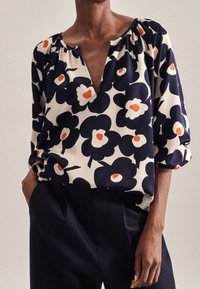 Blommig blus med djup V-ringning, korta puffärmar och en krämfärgad bakgrund. Har marinblå och orange blommönster, mjukt tyg.