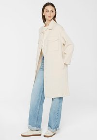 Beige Übergröße Mantel mit Kragen, frontaler Knopfleiste und einer Brusttasche, kombiniert mit hellblauen Jeans und cremefarbenen Sneakers.