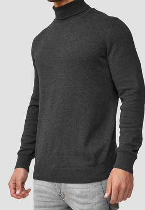 Homme portant un pull à col roulé gris charbon foncé et un jean gris clair, debout devant un fond uni gris clair.