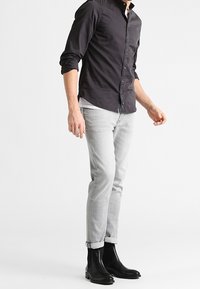 Camicia nera a bottoni, jeans slim-fit grigio chiaro e stivali alla caviglia in pelle nera. I dettagli includono maniche arrotolate e una trama liscia.