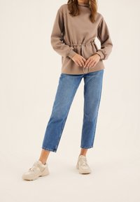 Świetlistobrązowy sweter z marszczonym pasem, długimi rękawami i sznurkiem. W połączeniu z niebieskimi, prostymi dżinsami z wysokim stanem i beżowymi trampkami.