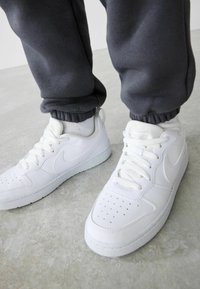 Hvide Nike sneakers med en glat læderoverdel, perforeret tåboks og tekstureret gummisål, parret med grå sweatpants med elastiske manchetter.