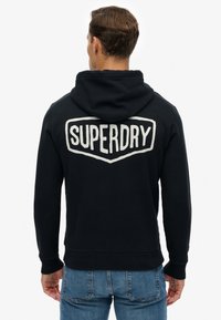 Sudadera negra con capucha que presenta un gran logo blanco "SUPERDRY" en la espalda. Confeccionada con tela suave y puños y dobladillo acanalados.