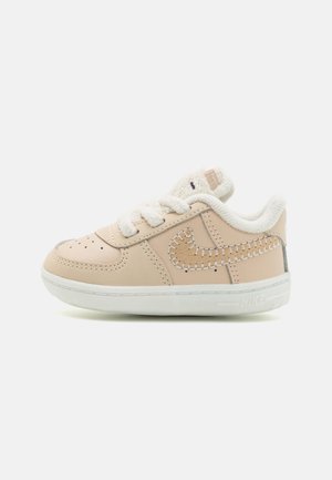 Beige toddler sneaker med hvid sål, syet lysebrun Nike swoosh, bløde hvide snørebånd og polstret krave mod hvid baggrund.