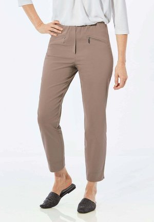 Hellbraune, schmal zulaufende Hose mit glatter Textur, ausgestattet mit zwei Reißverschluss-Vordertaschen. Getragen mit schwarzen Slippern.