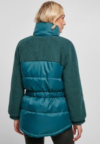Veste en polaire teal et résistante à l'eau, dotée d'un col haut, d'une ceinture élastique et de poignets côtelés. Le tissu est texturé et doux.