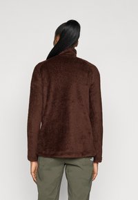 Casaco de fleece castanho com colarinho alto e superfície texturizada. Apresenta mangas longas e um corte descontraído, combinado com calças verde-oliva.