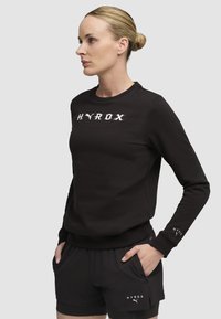 Čierna mikina s logom "HYROX" v bielej farbe, okrúhly výstrih, dlhé rukávy. V kombinácii s čiernymi šortkami s nápisom "HYROX" na leme.
