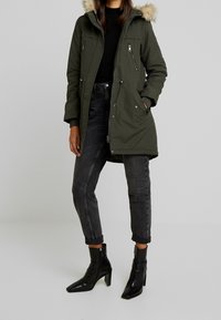 Olivgrön parkas med pälsfodrad huva, flera fickor med dragkedja och justerbar dragsko i midjan. Kombinerad med svarta högklackade ankellånga stövlar.