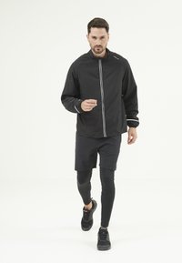 Svart löparjacka med reflexvita ränder, kombinerad med svarta shorts och leggings, bärs av en person i sportskor.