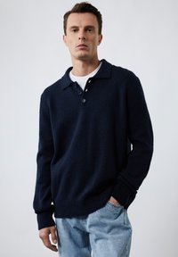 Marineblå strikket pullover med polo-krave, tre-knaps åbning, ribbede manchetter og kant; parret med lyseblå denim jeans.