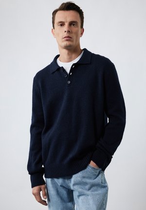 Navy gebreide pullover met een polo-kraag, drie knopen, geribbelde manchetten en onderrand; gecombineerd met lichtblauwe denim jeans.
