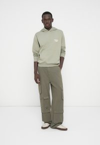 Νεαρός άνδρας με ανοιχτό πράσινο φούτερ με κουκούλα και το κείμενο «Originals Studio», ελιά καφέ παντελόνι cargo με πολλές τσέπες και άσπρα αθλητικά παπούτσια, σε χαλαρή στάση.