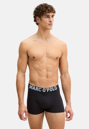 Homme torse nu avec des cheveux bouclés portant des boxer Marc O'Polo noirs, debout devant un fond clair uni, regardant sur le côté.