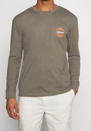 Homme portant une chemise à manches longues vert olive avec un logo globe orange et le texte "originals" sur la poitrine, associée à un pantalon blanc.