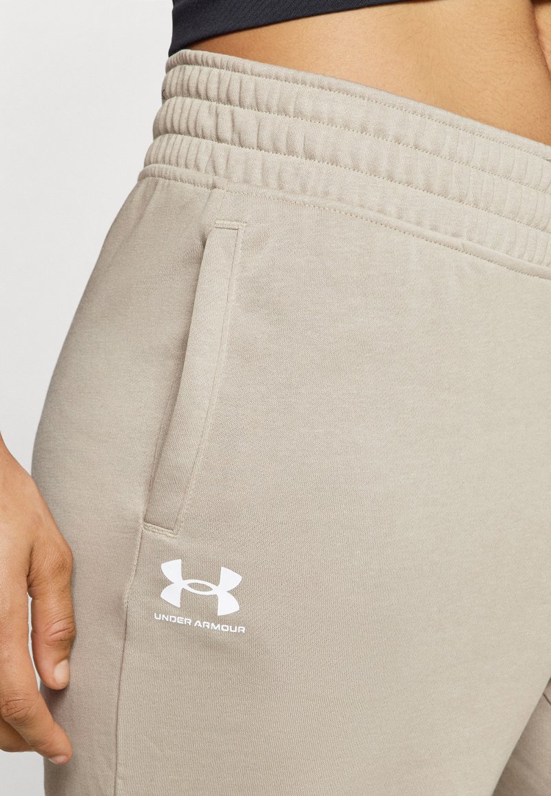 Helles beige Sporthose aus weichem Stoff, mit einem gerippten Bund und seitlichen Taschen. Ein weißes Under Armour-Logo ist prominent angebracht.