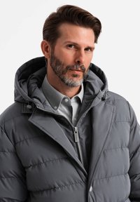 Giacca a vento grigia con cappuccio, caratterizzata da cuciture orizzontali, chiusura frontale con zip e un dettaglio con bottone. Indossata sopra una camicia colletto grigia chiara.