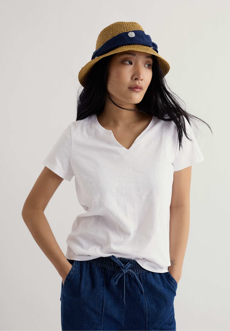 Femme portant un t-shirt blanc à col en V, un pantalon bleu à cordon de serrage et un chapeau de paille avec un ruban bleu marine, regardant sur le côté les mains dans les poches.