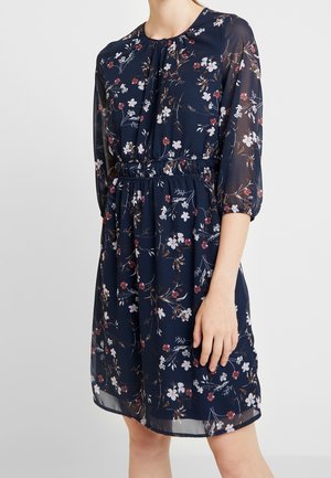 Robe bleu marine longueur genou avec taille froncée, manches longues en voile transparent, et motif floral blanc et rouge portée par une personne avec la main reposant sur le côté.