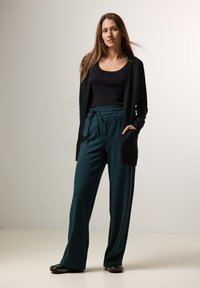 Svart cardigan med öppen front, matchad med turkosa vida byxor med elastisk midja. Svarta lägenheter med nitar.