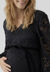 Melna mežģīņu grūtniecības kleita ar garām piedurknēm, V-veida kakla izgriezumu un pieguļošu siluetu, kuru akcentē samta josta ap vidukli.