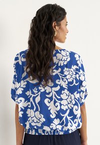 Blå bluse med hvidt blomstermønster, letvægtsstof, afslappet pasform, korte ærmer og rynket bundsøm, båret med ensfarvede marineblå bukser.