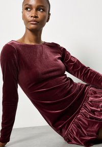 Bordowa sukienka z weluru z długimi rękawami, okrągłym dekoltem i marszczoną falbaną u dołu. Miękka faktura o gładkim wykończeniu.