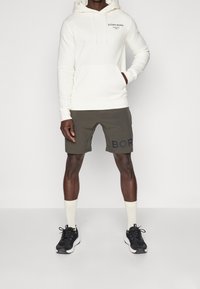 Weißer Hoodie mit Fronttasche und schwarzem Text, kombiniert mit olivgrünen Shorts mit "BORG" in dunkler Schrift. Schwarze Sneakers und weiße Socken.