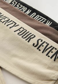 Shorts en tissu de coton dans des tons crème, marron et noir. Présentent de grands motifs de texte noir "VINGT QUATRE SEPT" et des coordonnées géographiques à la taille.