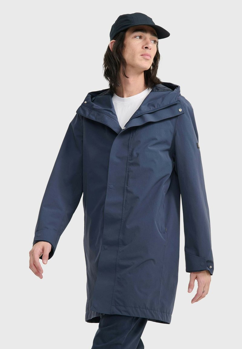 Manteau imperméable bleu marine avec capuche, fermeture à pression et deux poches latérales. Fabriqué en matériau lisse et léger, présentant une coupe décontractée.